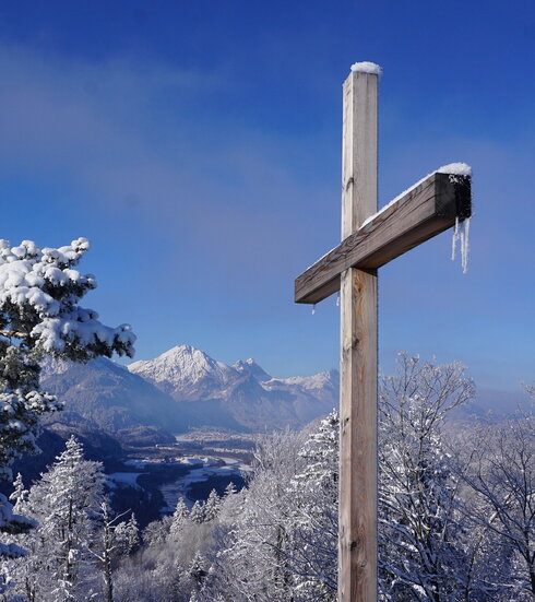 Auf dem Bild ist ein Gipfelkreuz mit Schnee und Eis bedeckt mit Bergen und Wald im Hintergrund zu sehen