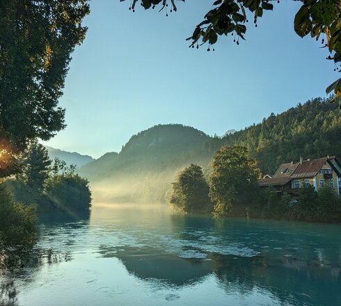 Das Bild fängt die ersten Sonnenstrahlen der Morgendämmerung am Lech ein, der sich dominant durch die Szene schlängelt. Der Fluss, ein tiefes Blau, liegt eingebettet in üppiges Grün, mit Bäumen und Büschen, die die Ufer säumen. Die ruhige Oberfläche des Flusses spiegelt die umliegende Landschaft wider. Rechts im Bild ist ein idyllisches Haus mit rotem Dach zu sehen. Obendrauf ist der Himmel ein helles Blau, durchzogen von ein paar Wolken. 