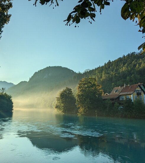 Das Bild fängt die ersten Sonnenstrahlen der Morgendämmerung am Lech ein, der sich dominant durch die Szene schlängelt. Der Fluss, ein tiefes Blau, liegt eingebettet in üppiges Grün, mit Bäumen und Büschen, die die Ufer säumen. Die ruhige Oberfläche des Flusses spiegelt die umliegende Landschaft wider. Rechts im Bild ist ein idyllisches Haus mit rotem Dach zu sehen. Obendrauf ist der Himmel ein helles Blau, durchzogen von ein paar Wolken. 