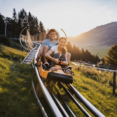 Das Bild zeigt eine Sommerrodelbahn mit einem Rodel in dem ein Junge und ein Mädchen sitzen. Im Hintergrund sieht man die Bergwelt und den Sonnenuntergang.