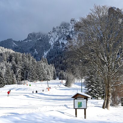 Das Bild zeigt einen hohen Berg. Auf einer Anhöhe davor ist ein Skilift samt Piste zu sehen. Zahlreiche Menschen stehen im Schlepplift und lassen sich nach oben fahren. Alles ist schneebedeckt. Der Himmel ist wolkenverhangen. Die Sonne scheint.  