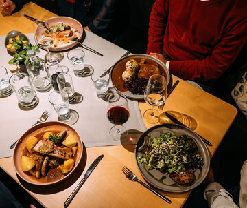 Menschen sitzen um einen Tisch mit Gerichten wie Knödeln, Fleisch und Salat. Es stehen Gläser, Besteck und kleine Flaschen auf dem Tisch.