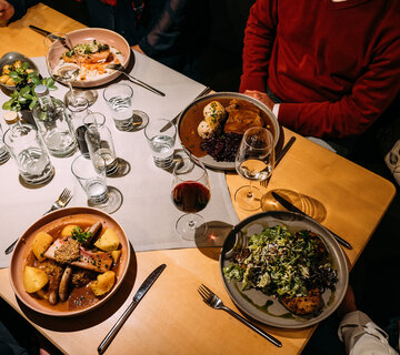 Menschen sitzen um einen Tisch mit Gerichten wie Knödeln, Fleisch und Salat. Es stehen Gläser, Besteck und kleine Flaschen auf dem Tisch.