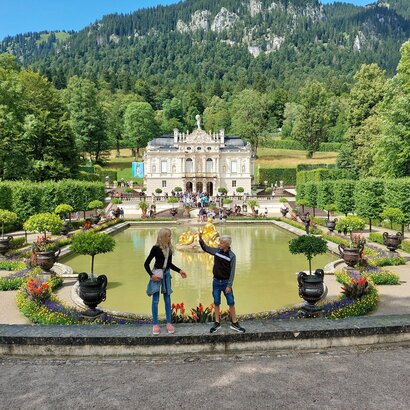  Das Bild fängt eine idyllische Szene in einem Park ein. Im Vordergrund stehen zwei Personen auf einer Steinmauer und blicken auf einen großen Teich. Der Teich ist von üppiger Vegetation umgeben, mit Bäumen und Sträuchern, die die Landschaft prägen. Ein Springbrunnen mit einer Statue befindet sich in der Mitte des Teichs und verleiht der Szene eine elegante Note. Im Hintergrund ragt ein majestätisches Burg-ähnliches Gebäude hoch, dessen Großzügigkeit durch die umgebende Vegetation betont wird. Der Himmel darüber ist ein klarer Blau, der auf einen angenehmen Tag hindeutet. Die Gesamtatmosphäre des Bildes ist eine der Ruhe und Schönheit.