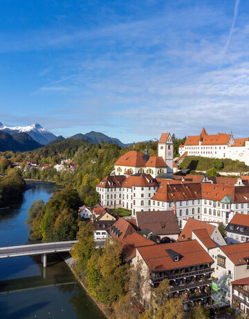 Das Foto zeigt das Hohen Schloss, dass auf einer Anhöhe über der Altstadt von Füssen thront. Direkt darunter ist der große Klosterkomplex des ehemaligen Benediktinerklosters mit seinen vielen Fenstern zu sehen. Alle Gebäude haben rote Dächer und weiße Wände. Neben den Gebäuden fließt der Lech, über den einen Brücke führt. Im Hintergrund erheben sich die Alpen, deren Gipfel schneebedeckt sind. Die Bäume haben bunte Blätter. Der Himmel ist ein wenig wolkenverhangen. 