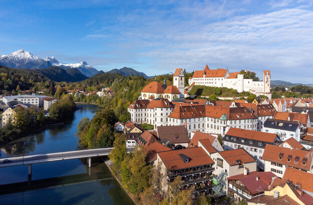 Das Foto zeigt das Hohen Schloss, dass auf einer Anhöhe über der Altstadt von Füssen thront. Direkt darunter ist der große Klosterkomplex des ehemaligen Benediktinerklosters mit seinen vielen Fenstern zu sehen. Alle Gebäude haben rote Dächer und weiße Wände. Neben den Gebäuden fließt der Lech, über den einen Brücke führt. Im Hintergrund erheben sich die Alpen, deren Gipfel schneebedeckt sind. Die Bäume haben bunte Blätter. Der Himmel ist ein wenig wolkenverhangen. 