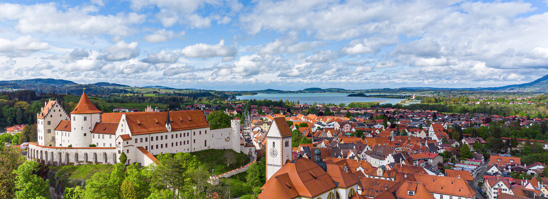 Das Bild zeigt die historische Altstadt Füssen mit dem Hohen Schloss, dem Klosterhof und dem Forggensee im Hintergrund. Der Himmel ist bewölkt.