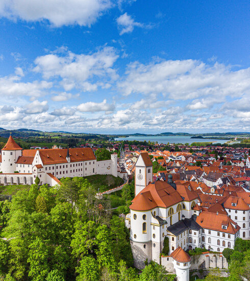 The picture offers a breathtaking view of the High Castle, the landmark of Füssen, Germany. The castle, a magnificent structure with a red roof, sits sublimely on a hill overlooking the town below. The town itself is a vibrant display of red roofs, creating an impressive contrast against the lush vegetation that surrounds it. The sky above is a clear blue, interspersed with fluffy white clouds that add to the overall serene and picturesque atmosphere of the scene. The perspective of the image is from a high view, providing a comprehensive view of the castle and the town below. The image conveys the essence of the castle and its surroundings, providing an insight into the rich history and architectural beauty of this famous landmark.