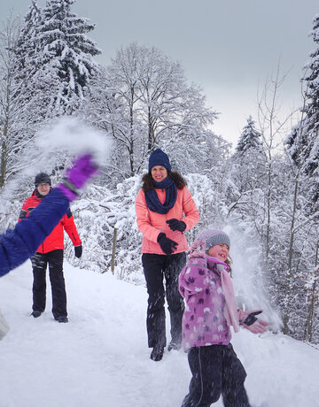 Das Bild fängt eine fröhliche Winterszene in einem schneebedeckten Wald ein. Eine Gruppe von Menschen, die in bunten Winterbekleidungen gekleidet sind, genießt den Schnee. Einige von ihnen werfen Schneebälle, was eine spielerische Atmosphäre schafft. Die Bäume im Hintergrund sind mit Schnee bedeckt, was zur winterlichen Atmosphäre beiträgt. Der Himmel über ihnen ist wolkenverhangen, was auf einen kalten und verschneiten Tag hinweist. Die Menschen stehen auf einem schneebedeckten Weg, der durch den Wald führt. Die gesamte Szene vermittelt ein Gefühl von Spaß und Kameradschaft, da die Gruppe von Freunden oder Familienmitgliedern Zeit miteinander in den Schnee verbringt.