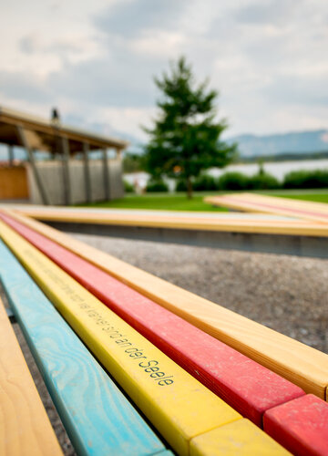 Das Bild zeigt zwei bunte Holzlatten, die in unterschiedlichen Winkeln aufeinander zulaufen. Auf einer gelben Holzlatte ist etwas eingraviert. Mann kann nur „sind an der Seele“ lesen. Im Hintergrund ist ein See zu sehen. Dahinter erheben sich hohe Berge. Der Himmel ist wolkenverhangen. 
