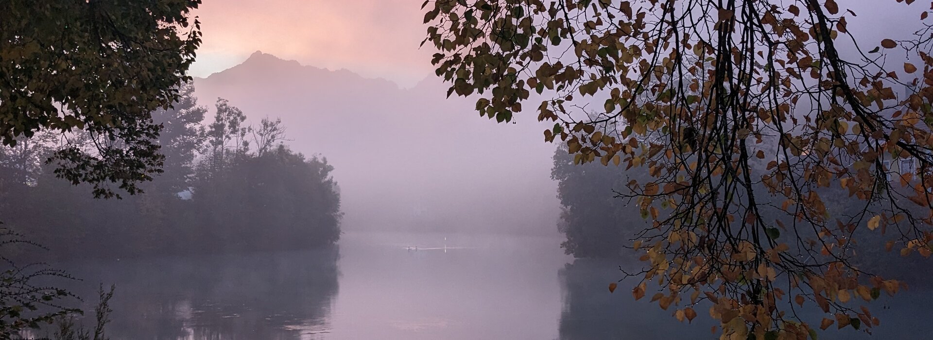 Das Bild zeigt eine friedliche Szene am Fluss Lech bei Sonnenaufgang. Der Himmel, der in Rosa und Lila-Töne getaucht ist, dient als atemberaubende Kulisse für das ruhige Wasser. Der Fluss, ruhig und unberührt, spiegelt die Farben des Himmels wider, was ein beeindruckendes Reflexionsbild erzeugt. Auf der linken und rechten Vorderseite des Bildes stehen Bäume, deren Äste in die Bildmitte reichen. Das Laub der Bäume, eine Mischung aus Grün und Gelb, bringen einen Hauch von Herbst in die Szene. 