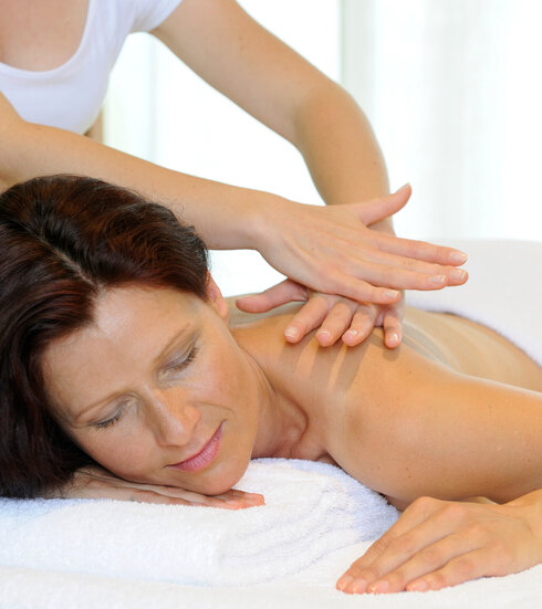 Das Bild zeigt eine Frau, die bäuchlings auf einer Massageliege liegt. Sie ist unbekleidet. Ein weißes Handtuch bedeckt ihr Gesäß und ihren unteren Rücken. Sie hat die Augen geschlossen. Über ihr steht eine Person, die ihr die obere Rückenpartie massiert. Die Person trägt ein weißes Oberteil. Der Raum ist hell erleuchtet.      