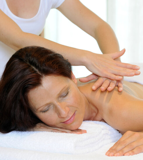 Das Bild zeigt eine Frau, die bäuchlings auf einer Massageliege liegt. Über ihr steht eine Person, die ihr die obere Rückenpartie massiert. 