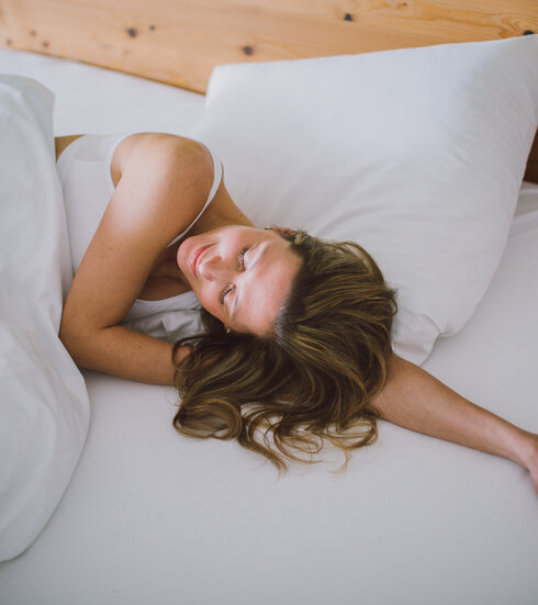 Das Bild zeigt eine Frau mit einem weißen kurzen Oberteil. Sie liegt in einem Bett, das weiß bezogen ist. Ihr Kopf liegt auf ihrem linken Arm. Sie hat die Augen geschlossen und wirkt entspannt. 