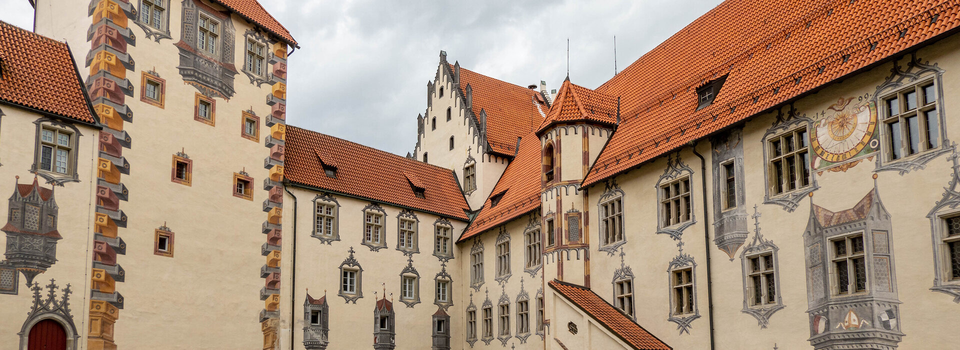 Das Bild zeigt den Innenhof des Hohen Schlosses in Füssen, der durch seine Illusionsmalerei an den Fassaden beeindruckt. Das Schlossgebäude hat rote Türen und ein Ziegeldach.