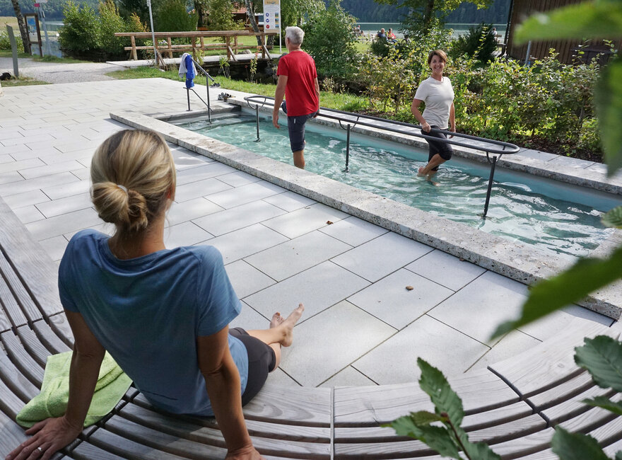 Das Bild zeigt zwei Personen, die in einem Tretbecken Wasser treten. Sie haben die Hosenbeine hochgekrempelt. Eine Person blickt auf eine Frau, die auf einer halbrunden Holzbank neben dem Tretbecken sitzt. Diese Frau hat dem Betrachter den Rücken zugedreht. Die Sonne scheint. 