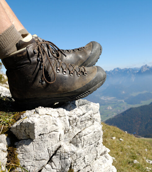 Das Bild zeigt eine Person, die auf einem Berg sitzt und nach unten blickt. Allerdings sind nur die ledernen Bergschuhe und die Wadern der Person zu sehen. Weiter unten erkennt man Bergwiesen, Wälder und ein Tal. Dahinter erheben sich weitere Berge. Der Himmel ist blau. Die Sonne scheint.  