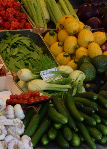 Dieses Bild zeigt einen Marktstand mit einer großen Auswahl an frischem Gemüse, darunter Tomaten, Zucchini, Auberginen, Gurken und Knoblauch. Die Farben sind lebendig und die Präsentation ist appetitlich, was die Frische der Produkte hervorhebt.