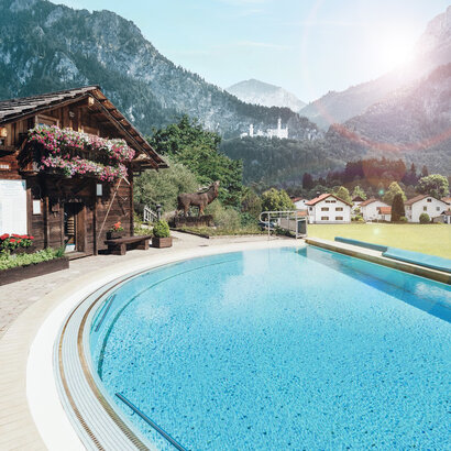 Das Bidl zeigt ein Außenbecken eines Schwimmbades. Im Hintergrund sieht man zwei Häuser und dahinter Berge und ein Schloss.