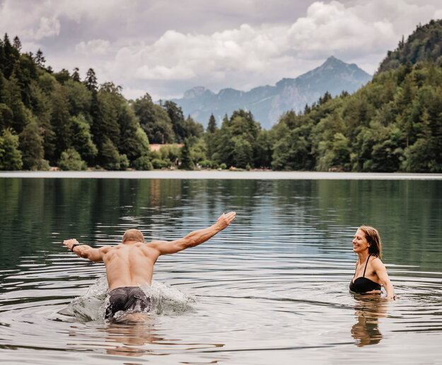 Das Bild zeigt einen Mann und eine Frau in Badekleidung in einem See. Der Mann macht einen Hechtsprung. Im Hintergrund sind die Berge und waldbedeckte Hänge zu erkennen.