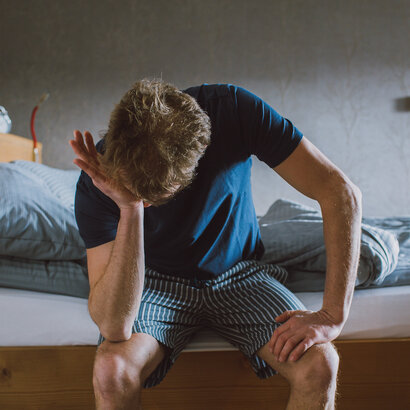 Das Bild zeigt einen Mann, der in einem Pyjama am Bettrand sitzt und die eine Hand auf den anderen Ellenbogen auf seine Knie gestützt hat. Mit der einen anderen Hand reibt er sich das Gesicht.