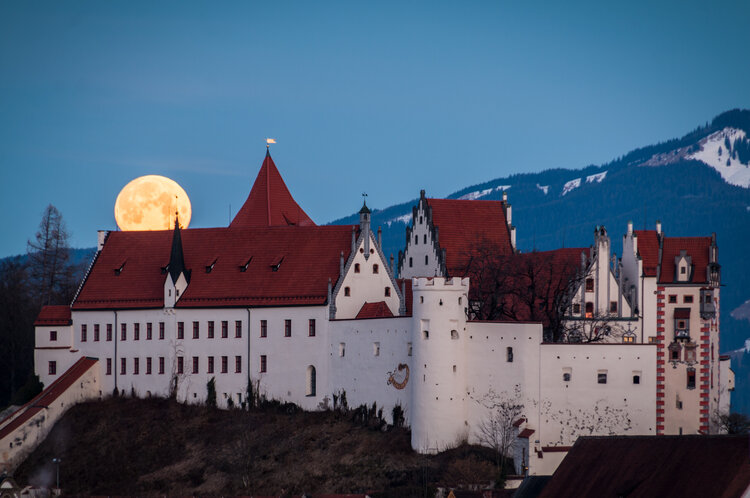 Das Hohe Schloss Füssen mit roten Dächern und Türmen vor einem großen, orangefarbener Vollmond. Schneebedeckte Berge im Hintergrund.