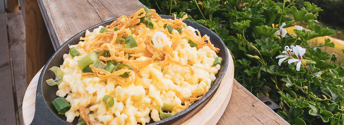 Ein Teller Käsespätzle in einer gusseisernen Pfanne wird auf einem Holztisch mit Blick auf eine atemberaubende Berglandschaft serviert. Die Szene vermittelt ein Gefühl von Gemütlichkeit und Genuss in den Bergen. 