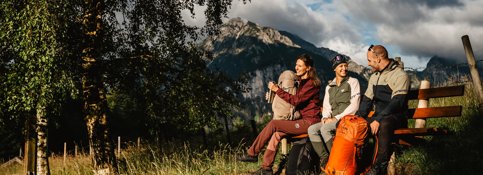 Das Bild zeigt zwei lachende Frauen und einen Mann in Outdoor-Kleidung, die auf einer Bank sitzen. Die Sonne scheint sie an und im Hintergrund sind die Allgäuer Alpen zu erkennen.