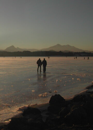 Das Bild zeigt zwei Menschen, die auf dem zugefrorenen Hopfensee in der Abendsonne spazieren