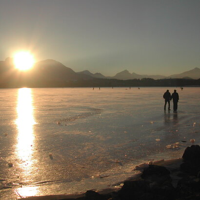 Das Bild zeigt zwei Menschen, die auf dem zugefrorenen Hopfensee in der Abendsonne spazieren