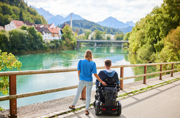 Das Bild fängt einen friedlichen Moment über einer Holzbrücke ein, die einen Blick auf eine Flusslandschaft bietet. Zwei Personen, ein Mann und eine Frau, stehen auf der Brücke, mit dem Rücken zur Kamera. Der Mann sitzt im Rollstuhl, während die Frau neben ihm steht. Beide sind lässig gekleidet, wobei die Frau ein blaues Shirt und der Mann einen schwarzen Mantel trägt. Die Brücke aus Holzdielen spannt sich über den Fluss und bietet einen Aussichtspunkt für den Betrachter. Der Fluss selbst ist ein lebendiges Blau, das den klaren Himmel darüber widerspiegelt. Im Hintergrund schmiegt sich ein malerisches Dorf in die üppige Vegetation, der das Bild mit Charme erfüllt. Die Atmosphäre des Bildes ist eine der Ruhe und Gesellschaft.