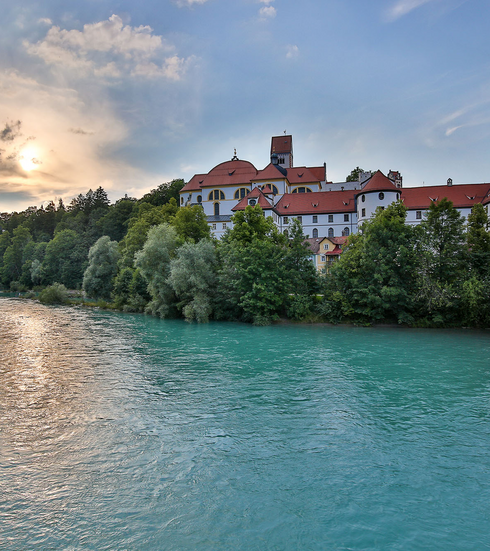 Das Bild zeigt ein schlossähnliches Gebäude, das auf einer Anhöhe am Ufer des Flusses Lech thront. Bei dem Gebäudekomplex handelt es sich um das ehemalige Benediktinerkloster St. Mang, das mit seinem roten Dach, majestätisch vor dem Hintergrund eines klaren blauen Himmels steht. Der türkise Fluss, fließt sanft vor dem Gebäude und spiegelt das Sonnenlicht. Die umgebende Landschaft ist üppig grün, mit Bäumen und Büschen, die die Landschaft prägen. Die untergehende Sonne tauch die Szene in warmes Licht.