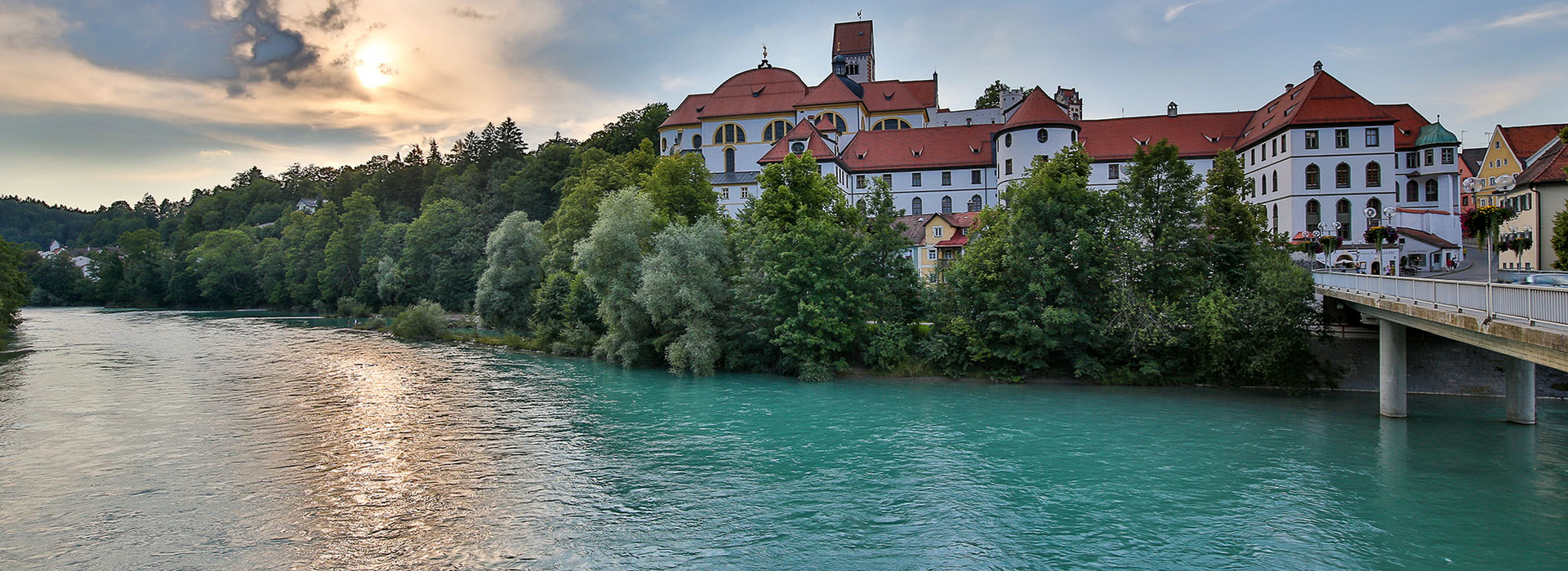 Das Bild zeigt ein schlossähnliches Gebäude, das auf einer Anhöhe am Ufer des Flusses Lech thront. Bei dem Gebäudekomplex handelt es sich um das ehemalige Benediktinerkloster St. Mang, das mit seinem roten Dach, majestätisch vor dem Hintergrund eines klaren blauen Himmels steht. Der türkise Fluss, fließt sanft vor dem Gebäude und spiegelt das Sonnenlicht. Die umgebende Landschaft ist üppig grün, mit Bäumen und Büschen, die die Landschaft prägen. Die untergehende Sonne tauch die Szene in warmes Licht.