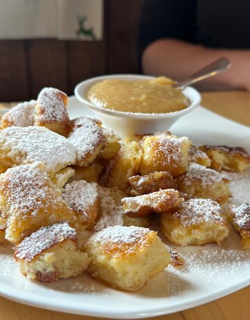Das Foto zeigt die Mehlspeise Kaiserschmarrn. Die mit weißem Puderzucker überzogen ist. Die Speise ist auf einem weißen Teller angerichtet. Ebenso auf dem Teller steht ein weißes Schälchen mit Apfelmus. 