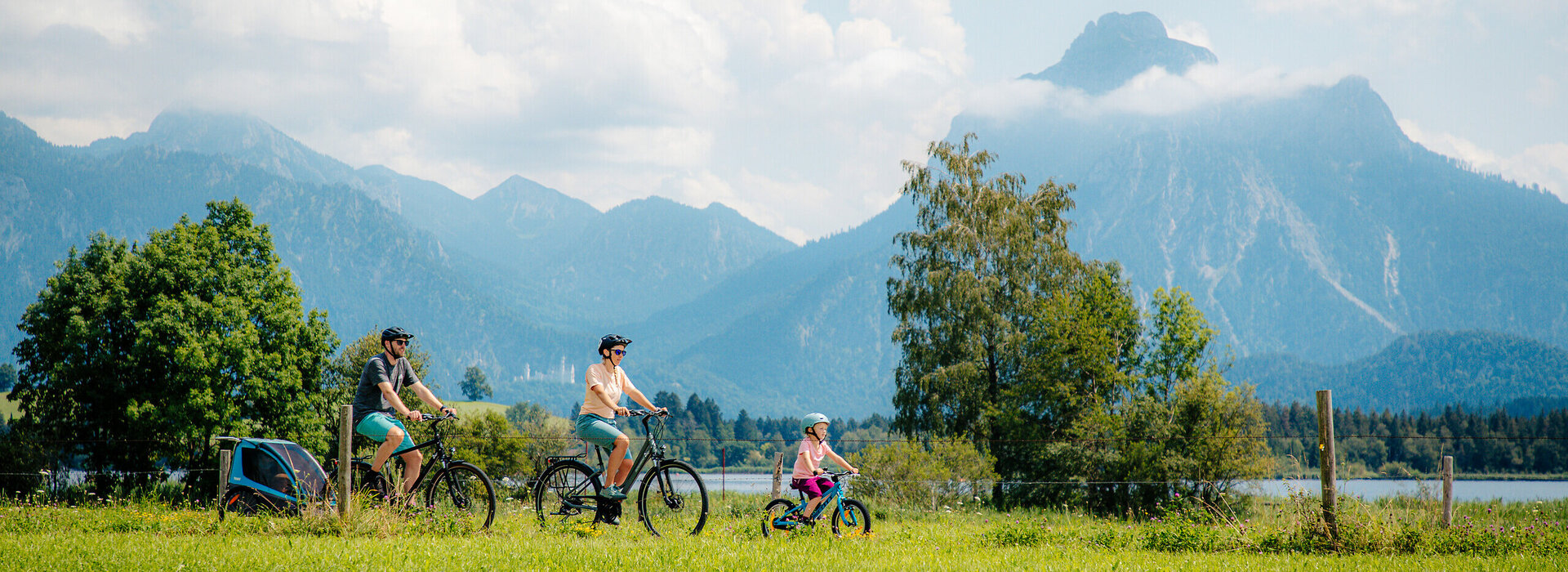 Das Bild fängt eine friedvolle Szene in einer grünen Wiese ein, wo drei Personen ein Fahrrad ausführen. Die Wiese ist üppig und grün, sie bietet eine lebendige Kulisse zur Szene. Der Himmel über ist ein klares Blau, garniert mit flauschigen weißen Wolken. In der Ferne erheben sich majestätische Berge gegen den Himmel, was der Szene ein Gefühl der Größe verleiht. Die Personen im Bild sind in lässiger Kleidung gekleidet, was auf einen gemütlichen Tag im Freien hinweist. Die Fahrräder, auf denen sie fahren, sind gut sichtbar, wobei eine Person ein Fahrrad reitet und die anderen beiden zu Fuß gehen. Die allgemeine Atmosphäre des Bildes ist die einer Ruhe und des Genusses der Natur.