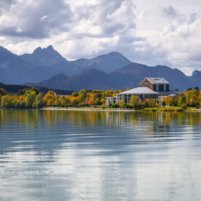 Das Foto wurde vom See aus aufgenommen. Am Ufer steht ein prächtiges Gebäude mit einem grauen Dach und vielen Fenstern. Der Mittelteil des Gebäudes ist rund. Es handelt sich um das Festspielhaus Neuschwanstein. Die Bäume ringsum haben schon eine bunte Farbe angenommen. Im Hintergrund erstreckt sich die Alpenkette. Der Himmel ist wolkenverhangen, trotzdem scheint die Sonne. 