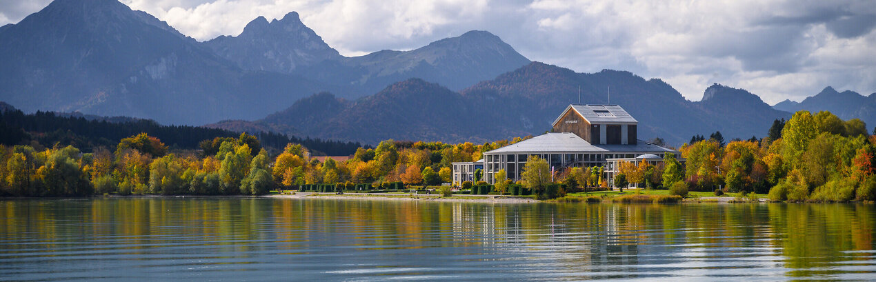 Das Foto wurde vom See aus aufgenommen. Am Ufer steht ein prächtiges Gebäude mit einem grauen Dach und vielen Fenstern. Der Mittelteil des Gebäudes ist rund. Es handelt sich um das Festspielhaus Neuschwanstein. Die Bäume ringsum haben schon eine bunte Farbe angenommen. Im Hintergrund erstreckt sich die Alpenkette. Der Himmel ist wolkenverhangen, trotzdem scheint die Sonne. 