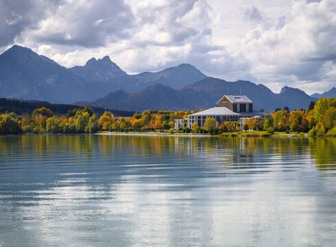 Das Foto wurde vom See aus aufgenommen. Am Ufer steht ein prächtiges Gebäude mit einem grauen Dach und vielen Fenstern. Der Mittelteil des Gebäudes ist rund. Es handelt sich um das Festspielhaus Neuschwanstein. Die Bäume ringsum haben schon eine bunte Farbe angenommen. Im Hintergrund erstreckt sich die Alpenkette. Der Himmel ist wolkenverhangen, trotzdem scheint die Sonne. 