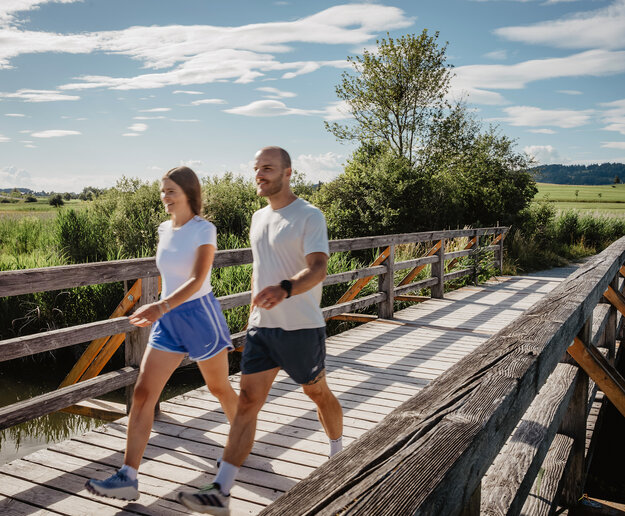Das Bild zeigt einen Mann und eine Frau, die über eine Brücke am Hopfensee walken