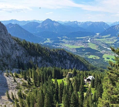 Das Bild zeigt eine weite Berglandschaft. Von oben ist eine große Berghütte von oben sowie noch weiter unten die Stadtgemeinde Reutte im Naturpark Tiroler Lech.