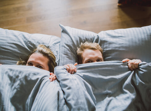 Das Bild zeigt eine Frau und einen Mann von oben, die sich eine blaugraue Bettdecke über die Nasenspitze ziehen und in die Kamera blicken. Ihre Köpfe sind auf ebenso blau-grauen Kissen gebettet.