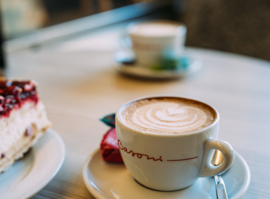 Ein Cappuccino mit kunstvoller Latte-Art steht auf einem Untertasse auf einem Tisch, neben einem Stück Kuchen.