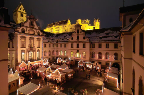 Blick auf die schneebedeckten Buden des Adventsmarkts Füssen im ehemaligen Kloster St. Mang und dem Hohen Schloss im Hintergrund bei Nacht