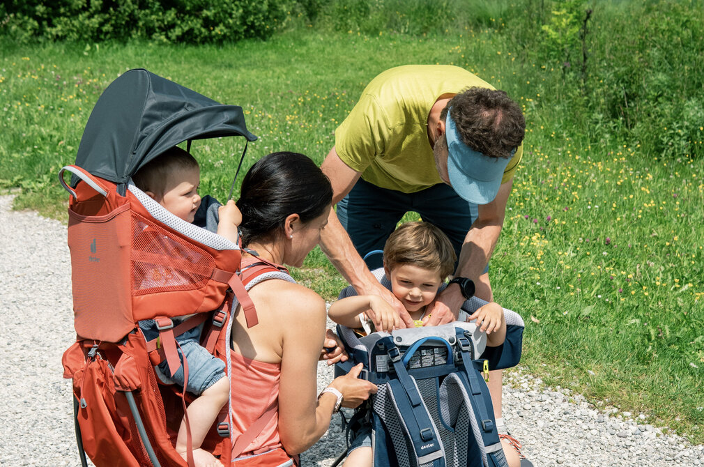 Eine Familie steht an einem Weg in der Natur. Eine Mutter sitzt mit einem Kleinkind im Tragesitz, während ein Vater mit einem älteren Kind an einem Rucksack arbeitet. Im Hintergrund ist ein See und eine grüne Landschaft zu sehen.