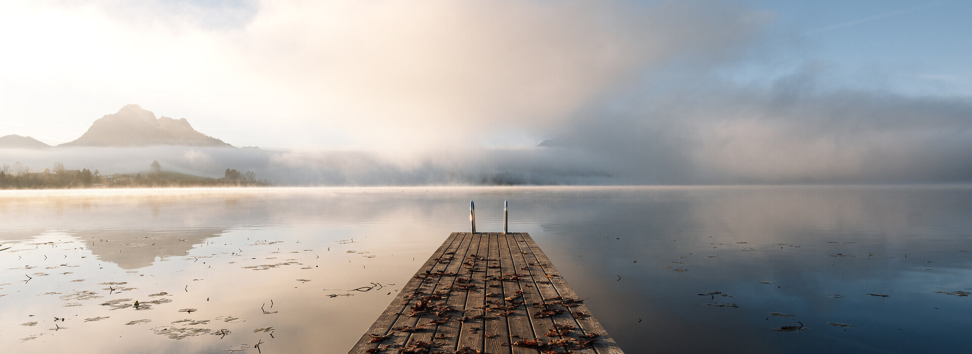 Das Bild zeigt einen Holzsteg, der den Hopfensee führt. Die Wasseroberfläche des Sees ist ruhig und fast wie ein Spiegel. Im Hintergrund erheben sich Berge aus dem Nebel. 