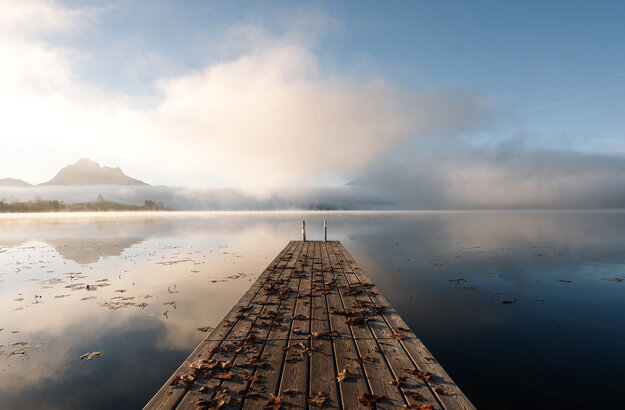 Das Bild zeigt einen Holzsteg, der den Hopfensee führt. Die Wasseroberfläche des Sees ist ruhig und fast wie ein Spiegel. Im Hintergrund erheben sich Berge aus dem Nebel. 