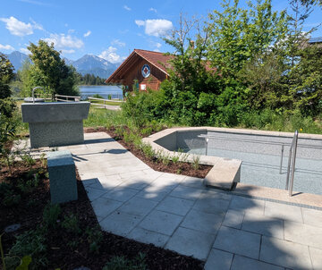 Kneipp-Anlage_Hopfen am See Ein neu gestalteter Außenbereich mit einem flachen Pool und einem Handlauf. Natursteinplatten bilden den Gehweg, umgeben von Büschen und kleinen Pflanzen. Im Hintergrund ist ein Wasserlauf sowie ein Holzgebäude sichtbar, das für Erholung genutzt werden kann. Die Landschaft ist von Bergen umgeben.