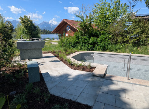 Ein neu gestalteter Außenbereich mit einem flachen Pool und einem Handlauf. Natursteinplatten bilden den Gehweg, umgeben von Büschen und kleinen Pflanzen. Im Hintergrund ist ein Wasserlauf sowie ein Holzgebäude sichtbar, das für Erholung genutzt werden kann. Die Landschaft ist von Bergen umgeben.