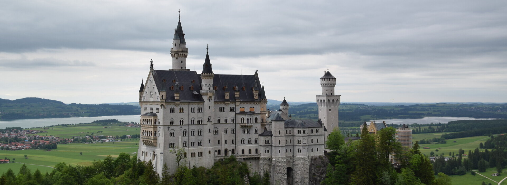 Das Bild zeigt das weiße Schloss Neuschwanstein, dass auf einer bewaldeten Anhöhe vor einer weiten Ebene liegt. Auf der Ebene sind Seen, Ortschaften und grüne Wiesen zu erkennen. Das Schloss Neuschwanstein steht im Fokus des Fotos. Es besteht aus mehreren unterschiedlichen Gebäudeteilen, wie Türmen, Erkern und einem Torbau, die miteinander verbunden sind. Das Hauptgebäude des Schlosses ist 5-stöckig. Es hat ein graues Dach. Auf dem Hauptgebäude stehen eine überlebensgroße Statue sowie eine Löwenfigur. Der Himmel ist grau und wolkenverhangen. 