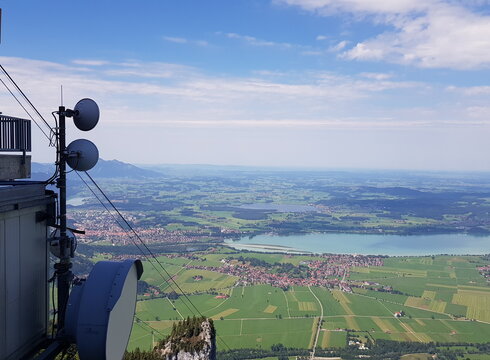 Das Foto zeigt das Voralpenland von der Bergstation der Tegelbergbahn aus. Man sieht unterschiedlichen Ortschaften, die um einen großen See angesiedelt sind. Man blickt in die weite Ebene, die mit Wäldern, Wiesen und weiteren vereinzelten kleinen Ortschaften durchzogen ist.  Im Vordergrund sieht man einen Teil der Bergstation  und die Tragseile der Bergbahn die in die Tiefe führen. 
